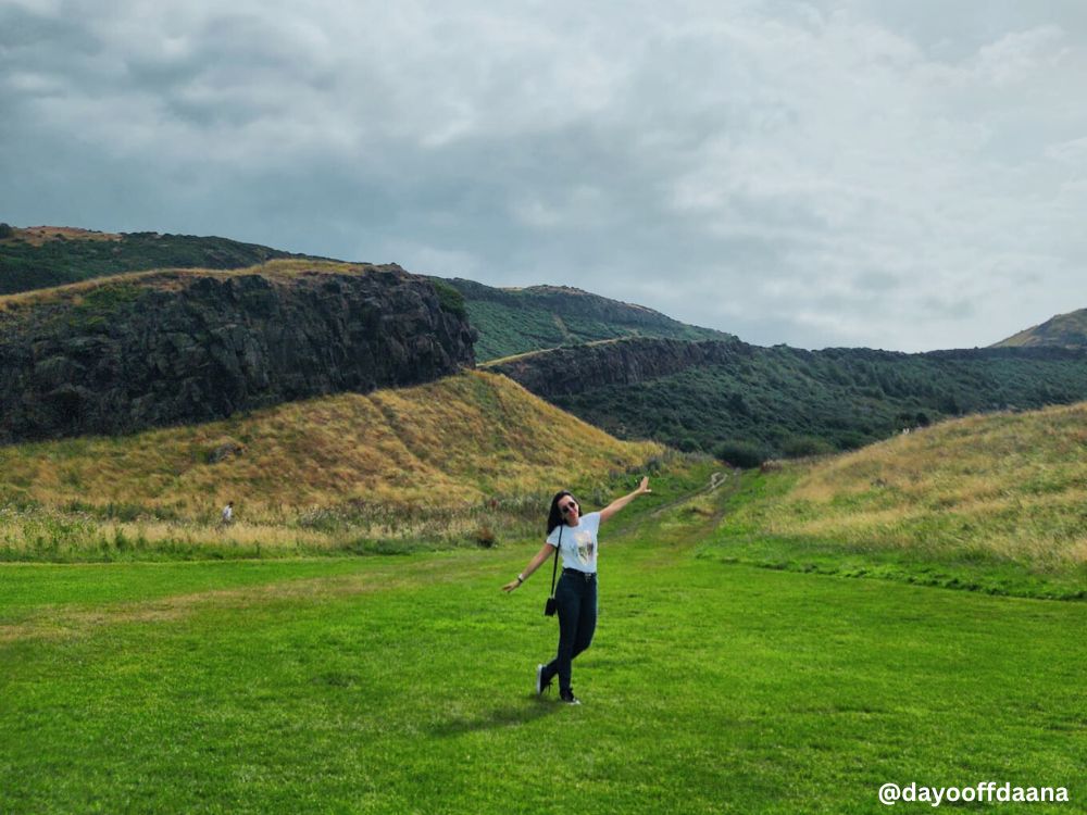 O-que-fazer-em-Edimburgo-Arthur- Seat O que fazer em Edimburgo: a foto mostra a montanha na natureza conhecida como Arthur’s Seat, e eu bem no meio da paisagem