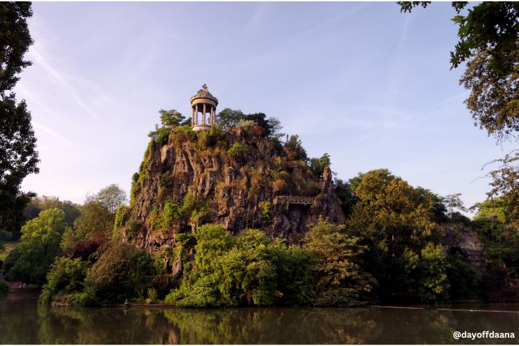 Alt: Foto mostrando um ponto do Parc des Buttes-Chaumont, com o templo inspirado em Tivoli, na Itália, que oferece uma vista panorâmica da cidade.